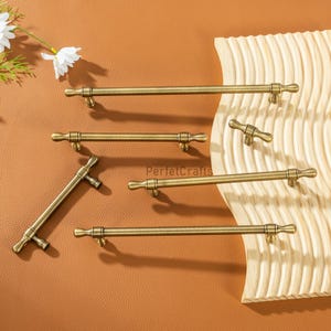 May include: A selection of antique brass cabinet pulls and handles are arranged on a brown surface. The handles vary in size and orientation, highlighting their design and finish. The hardware has a classic, vintage aesthetic.