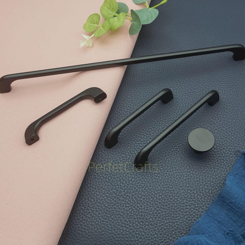 Op de afbeelding: Een verzameling zwarte meubelbeslag, waaronder lange stanggrepen, gebogen handgrepen en een ronde knop. De hardware wordt gepresenteerd op een roze en blauw oppervlak, met een takje groen op de achtergrond.