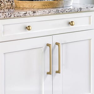 May include: White kitchen cabinets with gold-colored hardware. The upper cabinets have round knobs, while the lower cabinets feature long, rectangular pulls. The countertop is a speckled granite.