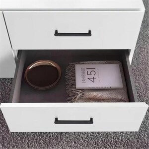 May include: White bedside cabinet with two drawers, one open. The open drawer reveals a brown tray, a book titled "Fahrenheit 451", and a beige fringed blanket. Black handles are on the drawers.