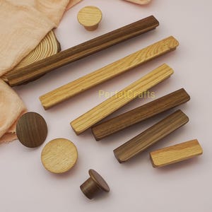 May include: A collection of wooden cabinet knobs and pulls in various shades of brown and tan. The knobs are round, and the pulls are rectangular. The text "PerfectCrafts" is visible on the image. The items are arranged on a light pink surface.