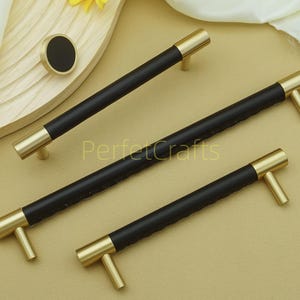 Puede incluir: Un juego de tiradores de armario negros y dorados y un pomo a juego. Los tiradores tienen un centro negro texturizado con extremos y tiradores dorados. El pomo es redondo con un centro negro y ribete dorado. La palabra "PerfectCrafts" es visible.