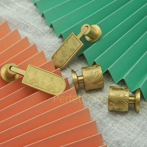 May include: Close-up of several brass cabinet knobs and pulls. The knobs have a cylindrical base and a decorative, swirling pattern. The pulls have a rectangular face with a wave-like design. The hardware is displayed on a folded paper background.