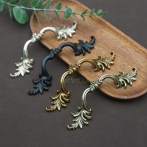 May include: An assortment of decorative cabinet handles in silver, black, gold, and antique brass finishes. Each handle features a curved design with ornate leaf detailing, perfect for adding a touch of elegance to furniture. The handles are displayed on a wooden tray.