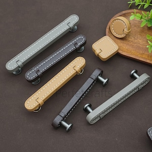 May include: A collection of cabinet pulls and knobs in various colors, including gray, brown, and tan. The pulls have a leather-like appearance with white stitching and metal hardware. A round and square knob are also included.