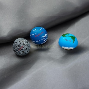 May include: Three spherical decorative objects: a gray moon with craters and the word "Moon", a blue Neptune with white lines and the word "Neptune", and a blue Earth with green continents and the word "Earth".