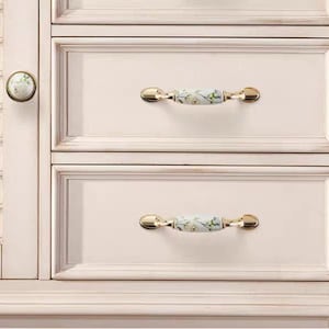 Pu&ograve; includere: Primo piano di un mobile color crema con due cassetti. I cassetti hanno maniglie dorate con un motivo floreale. Un pomello rotondo con un motivo floreale &egrave; sulla porta dell'armadio.