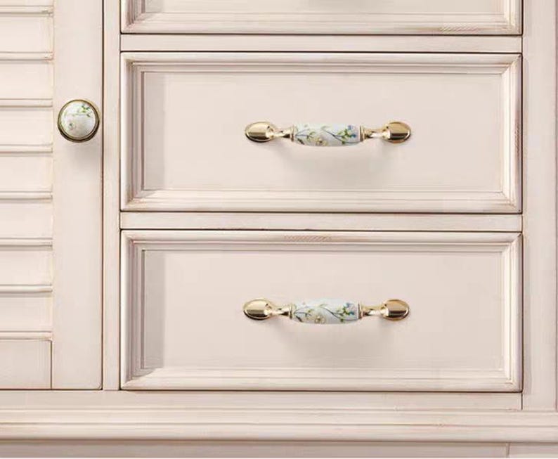 Pu&ograve; includere: Primo piano di un mobile color crema con due cassetti. I cassetti hanno maniglie dorate con un motivo floreale. Un pomello rotondo con un motivo floreale &egrave; sulla porta dell'armadio.