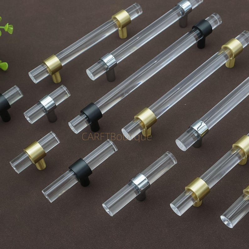 May include: Clear acrylic cabinet handles with gold, silver, and black metal accents. The handles are cylindrical and arranged on a brown surface. The hardware is designed for furniture and home decor.