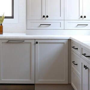 May include: A modern kitchen with white cabinetry and a light gray countertop. The cabinets have black hardware, including knobs and bar pulls. A window is visible on the left, and a vase with dried plants sits on the countertop.