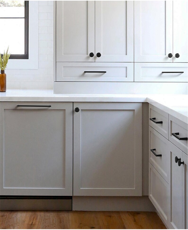 May include: A modern kitchen with white cabinetry and a light gray countertop. The cabinets have black hardware, including knobs and bar pulls. A window is visible on the left, and a vase with dried plants sits on the countertop.