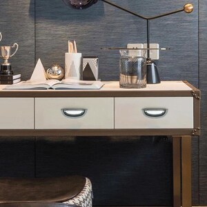 Może przedstawiać: Kremowe biurko z trzema szufladami, każda z srebrnym uchwytem w kształcie litery D. Biurko ma brązową ramę i złote metalowe nogi. Na blacie biurka znajdują się r&oacute;żne przedmioty dekoracyjne, w tym trofeum, szklany wazon i lampa.