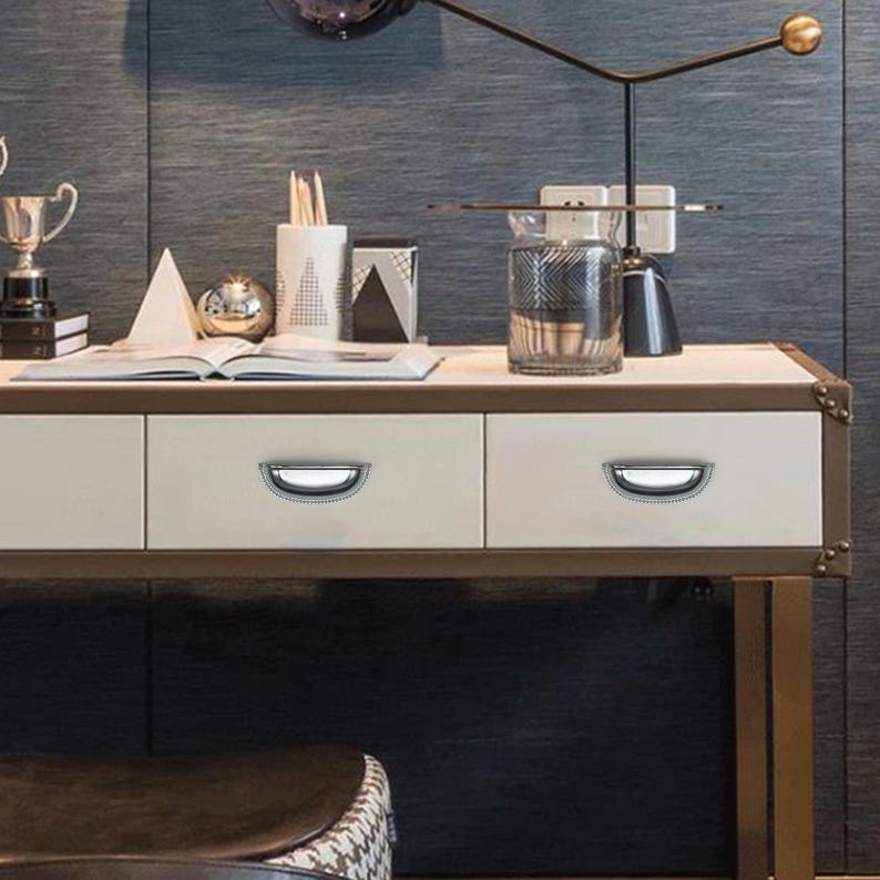 Może przedstawiać: Kremowe biurko z trzema szufladami, każda z srebrnym uchwytem w kształcie litery D. Biurko ma brązową ramę i złote metalowe nogi. Na blacie biurka znajdują się r&oacute;żne przedmioty dekoracyjne, w tym trofeum, szklany wazon i lampa.