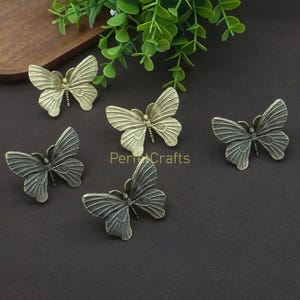 May include: Five decorative butterfly-shaped hardware pieces in varying shades of bronze and gray. The butterfly wings are detailed with vein patterns. The hardware is displayed on a dark surface with greenery in the background.