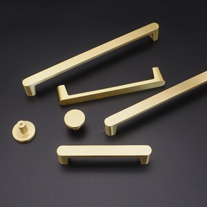 May include: A selection of gold-coloured cabinet hardware, including handles and a knob. The hardware has a sleek, modern design with rounded edges. The pieces are arranged on a dark background, highlighting their metallic finish.