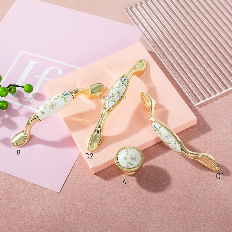 Pu&ograve; includere: Una collezione di ferramenta per mobili color oro, tra cui maniglie e un pomello. Le maniglie presentano un centro in ceramica bianca con un motivo floreale. La ferramenta &egrave; esposta su una superficie rosa, mettendo in risalto gli elementi decorativi.