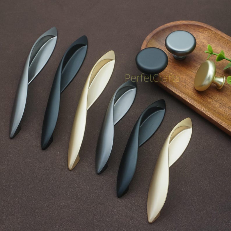 以下が含まれることがあります： さまざまな金属仕上げのキャビネットハードウェアのコレクション。シルバー、ブラック、ゴールドのカーブしたハンドルと、お揃いの色の丸いノブが含まれています。ハードウェアは茶色の表面に表示されています。