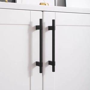 K&ouml;nnte beinhalten: Nahaufnahme eines wei&szlig;en Schranks mit zwei schwarzen, strukturierten T-Stangen-Griffen. Die Griffe sind vertikal angebracht und an den Schrankt&uuml;ren befestigt. Das Design ist minimalistisch und modern.