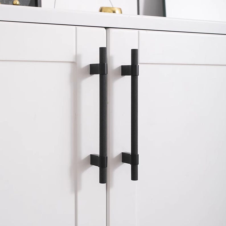 K&ouml;nnte beinhalten: Nahaufnahme eines wei&szlig;en Schranks mit zwei schwarzen, strukturierten T-Stangen-Griffen. Die Griffe sind vertikal angebracht und an den Schrankt&uuml;ren befestigt. Das Design ist minimalistisch und modern.