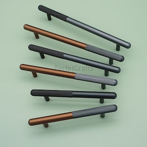 Könnte beinhalten: Eine Auswahl von sechs Schrankgriffen in verschiedenen Farbkombinationen. Jeder Griff hat ein zylindrisches Design mit einem geriffelten Abschnitt und einem glatten, abgerundeten Griff. Die Farbschemata umfassen Schwarz, Bronze und Grau und bieten eine moderne Ästhetik.