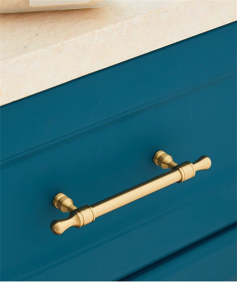 May include: A gold-coloured cabinet pull handle with a bar and rounded ends, mounted on a teal blue cabinet drawer. The handle is centred horizontally, adding elegance to the cabinet.