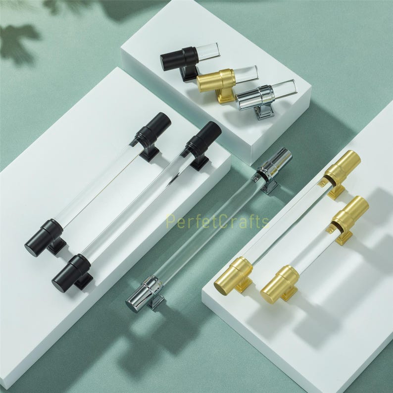 May include: Cabinet hardware featuring clear acrylic rods with black, gold, and silver metal accents. The handles are displayed on white platforms against a light green background. The text "PerfectCrafts" is visible.