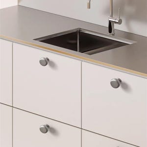 May include: A modern kitchen sink with a stainless steel countertop and a chrome faucet. The sink is square and set into the countertop. White cabinets with silver knobs are below the countertop.