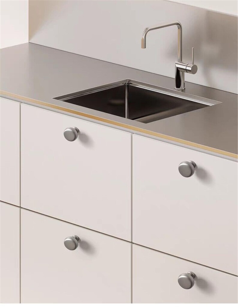 May include: A modern kitchen sink with a stainless steel countertop and a chrome faucet. The sink is square and set into the countertop. White cabinets with silver knobs are below the countertop.