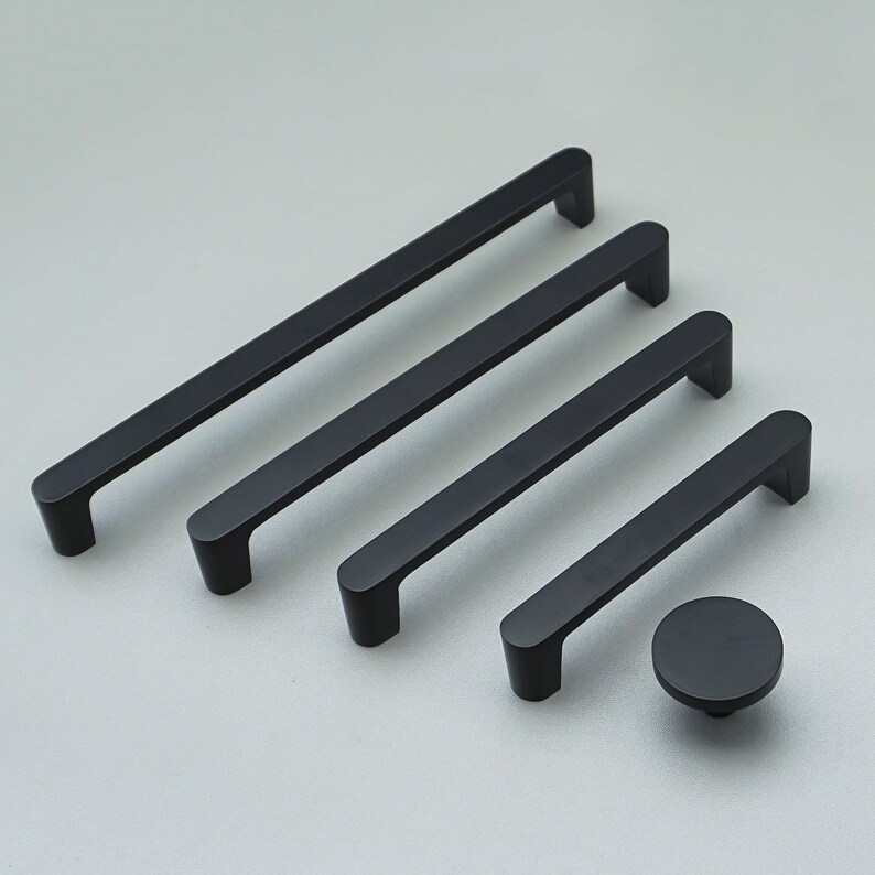 May include: A set of matte black cabinet handles and a knob. The set includes four rectangular bar pulls of different lengths and a circular knob. The hardware is displayed on a light grey surface, showcasing a modern design.