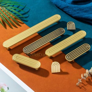 May include: A collection of gold and black cabinet hardware, including handles and knobs. The handles have a grooved design, and the knobs are arched. The hardware is arranged on a surface with orange, blue, and green colors.