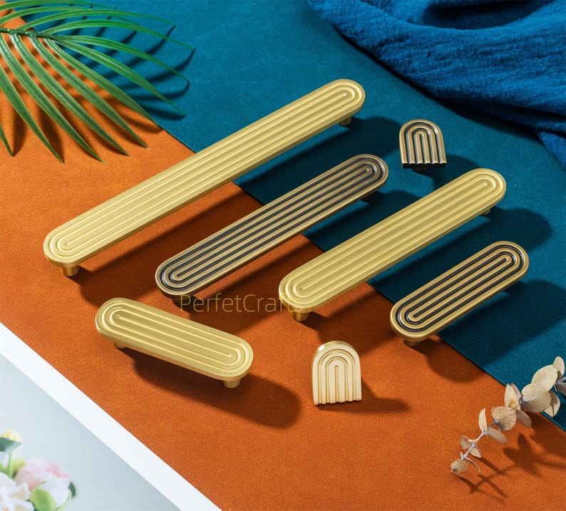 May include: A collection of gold and black cabinet hardware, including handles and knobs. The handles have a grooved design, and the knobs are arched. The hardware is arranged on a surface with orange, blue, and green colors.