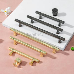 May include: An assortment of cabinet hardware, including gold and black textured bar pulls and matching knobs. The hardware is displayed on a white surface with a pink background. The text "PerfetcCrafts" is visible.