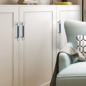 Puede incluir: Puertas de armario blancas con tiradores plateados y azules. Los tiradores tienen un centro cil&iacute;ndrico azul y extremos plateados. Un sill&oacute;n azul claro con un coj&iacute;n estampado y una manta gris en el fondo.