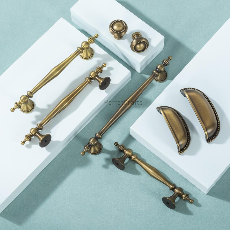 Może przedstawiać: Asortyment antycznych mosiężnych okuć do szafek, w tym uchwyty, gałki i uchwyty do kubk&oacute;w. Okucia mają wygląd vintage z ozdobnymi detalami i ciepłym, złotym odcieniem. Elementy są ułożone na białej powierzchni.