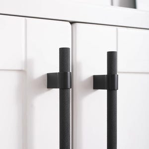 K&ouml;nnte beinhalten: Nahaufnahme von zwei schwarzen Schrankgriffen mit strukturiertem, zylindrischem Design. Die Griffe sind an wei&szlig;en Schrankt&uuml;ren montiert. Die Hardware hat eine moderne, minimalistische &Auml;sthetik, die f&uuml;r K&uuml;chen- oder Badezimmerschr&auml;nke geeignet ist.