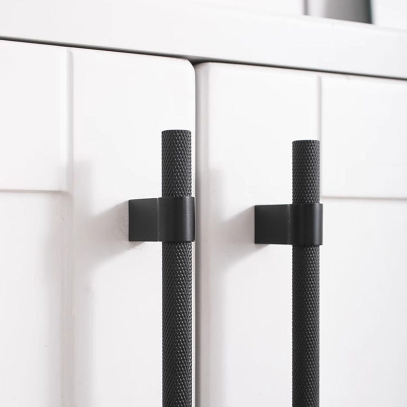 K&ouml;nnte beinhalten: Nahaufnahme von zwei schwarzen Schrankgriffen mit strukturiertem, zylindrischem Design. Die Griffe sind an wei&szlig;en Schrankt&uuml;ren montiert. Die Hardware hat eine moderne, minimalistische &Auml;sthetik, die f&uuml;r K&uuml;chen- oder Badezimmerschr&auml;nke geeignet ist.