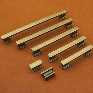 Puede incluir: Juego de tiradores y pomos de armario de latón antiguo. La ferretería presenta un diseño rectangular con un acabado en bronce oscuro. Los tiradores son de varias longitudes y los pomos son pequeños y cuadrados.