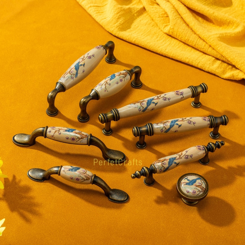 May include: A collection of antique-style cabinet hardware, including handles and a knob. The hardware features a cream-colored ceramic center with a blue bird and floral design, set against a bronze-toned metal base. The text "PerfectCrafts" is visible.