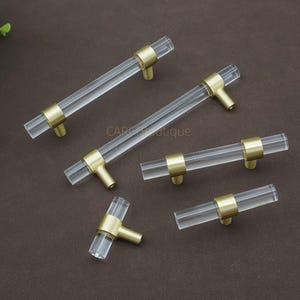 May include: A selection of clear acrylic cabinet pulls with brass-coloured accents. The pulls have a cylindrical design with brass rings at each end and a brass stem. The hardware is arranged on a brown surface.