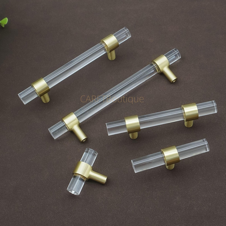 May include: A selection of clear acrylic cabinet pulls with brass-coloured accents. The pulls have a cylindrical design with brass rings at each end and a brass stem. The hardware is arranged on a brown surface.