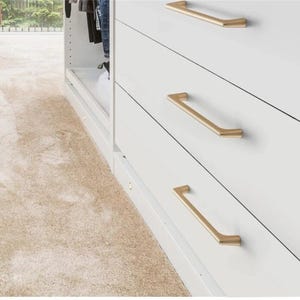 May include: A close-up of a white dresser with three drawers, each featuring a gold-colored, angular handle. The handles are horizontally aligned. The dresser is part of a larger closet system, with a beige carpet in the foreground.