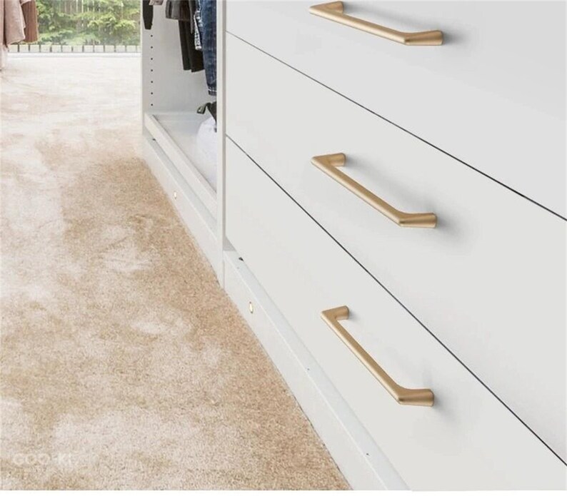 May include: A close-up of a white dresser with three drawers, each featuring a gold-colored, angular handle. The handles are horizontally aligned. The dresser is part of a larger closet system, with a beige carpet in the foreground.