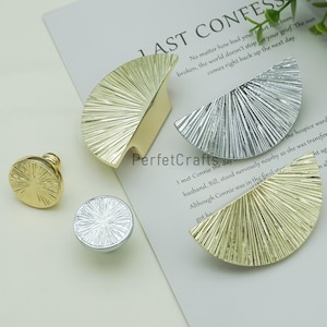 May include: A collection of decorative hardware, including gold and silver cabinet knobs and drawer pulls. The knobs are round with a sunburst design, while the pulls are semi-circular with a similar textured pattern. The hardware is displayed on a white surface.