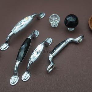 Peut inclure: Ensemble de poignées et boutons de meubles. Les poignées sont en métal argenté avec des accents en cristal clair et noir facetté. Deux boutons en cristal sont également présents. Présentation sur fond marron, divers modèles.