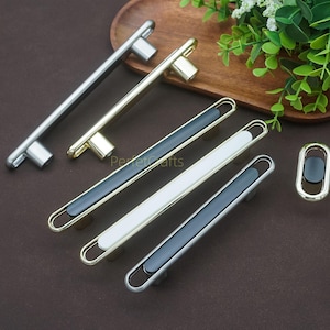 May include: A selection of contemporary cabinet handles in different styles and finishes. The handles feature silver, gold, and black and white details. The hardware is presented on a brown surface with greenery in the background.