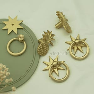 May include: A selection of gold-toned cabinet hardware, featuring pineapple-shaped knobs, starburst ring pulls, and a star-shaped backplate. The hardware is presented on a pale green surface, possibly for a tropical or nautical design.