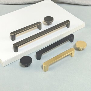 May include: A selection of cabinet handles and knobs in black, gold, and bronze finishes. The handles are rectangular, and the knobs are circular. The hardware is presented on a white surface, suitable for kitchen or furniture projects.