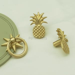 May include: Gold-toned cabinet hardware, including a pineapple-shaped knob, a starburst pull with a ring handle, and a pineapple knob. The hardware has a textured finish and is set against a light background. The text "Perfectcrafts" is visible.