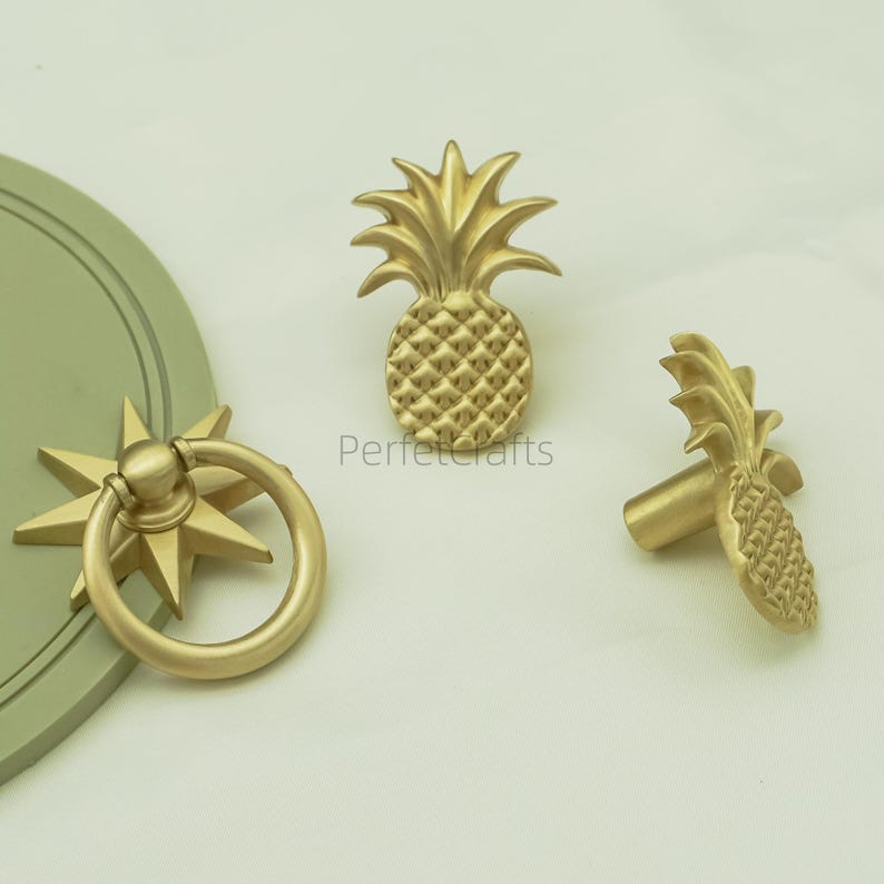 May include: Gold-toned cabinet hardware, including a pineapple-shaped knob, a starburst pull with a ring handle, and a pineapple knob. The hardware has a textured finish and is set against a light background. The text "Perfectcrafts" is visible.