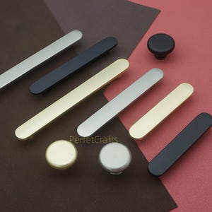 May include: A collection of cabinet hardware, including silver, black, and gold-toned bar pulls and round knobs. The hardware is displayed on a brown and red surface, showcasing various finishes and styles for home decor.